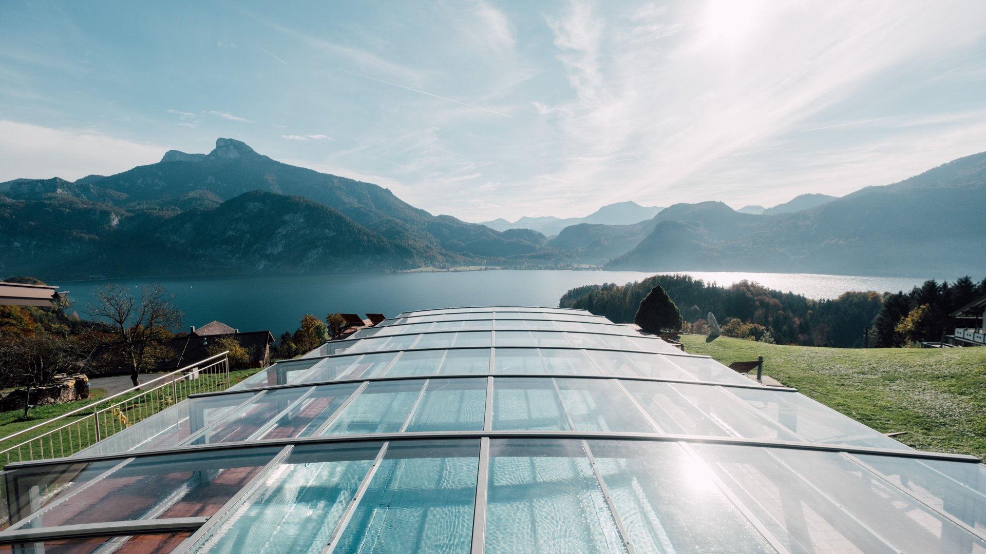 Panoramablick auf den Mondsee Blick auf einen See umgeben von Bergen mit einem gläsernen Außenpool im Vordergrund.